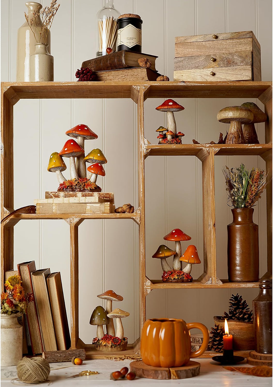 A rustic wooden display shelf styled with decorative mushrooms, books, dried flowers, a small wooden box, a ceramic pumpkin mug, and candles against a white paneled wall.