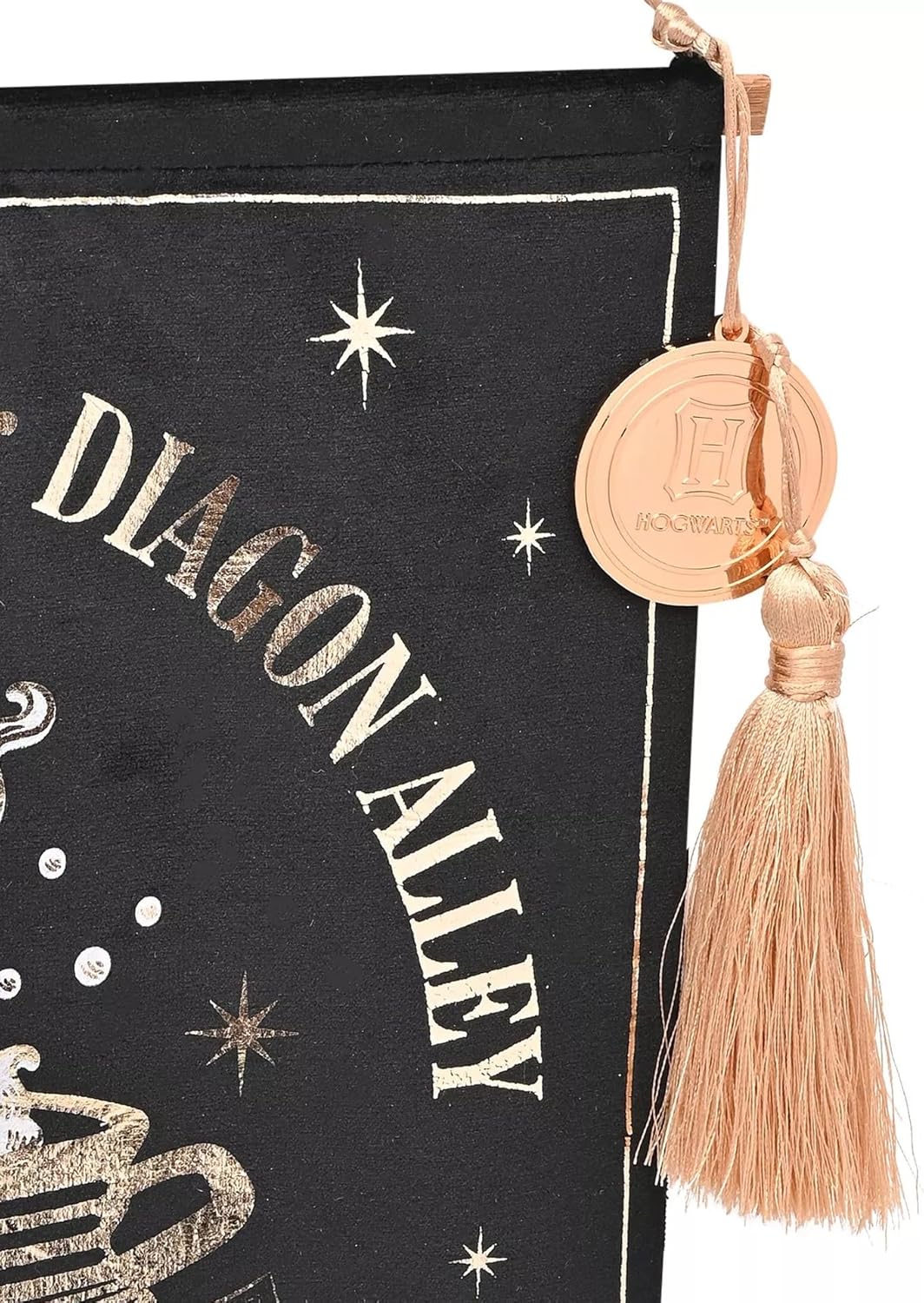 A close-up of a black fabric hanging pouch or bookmark with metallic gold foil text reading 'DIAGON ALLEY' and star motifs, finished with a tassel and round gold charm engraved with 'H Nourrit'.