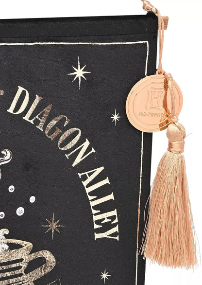 A close-up of a black fabric hanging pouch or bookmark with metallic gold foil text reading 'DIAGON ALLEY' and star motifs, finished with a tassel and round gold charm engraved with 'H Nourrit'.