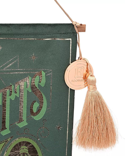 A close-up of a dark green decorative hanging piece with metallic gold and green lettering, a hanging cord, a round gold coin charm, and a tassel.