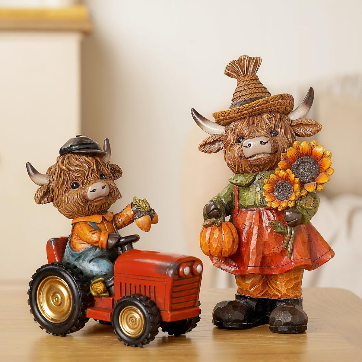Two decorative cow figurines, one on a red tractor and the other holding pumpkins and sunflowers, on a light background.
