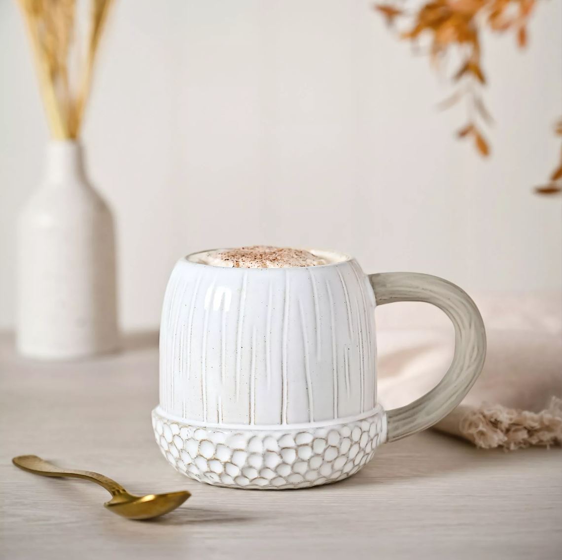 White textured mug with a handle on a light surface, accompanied by a gold spoon and decorative elements.
