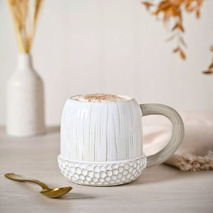 White textured mug with a handle on a light surface, accompanied by a gold spoon and decorative elements.