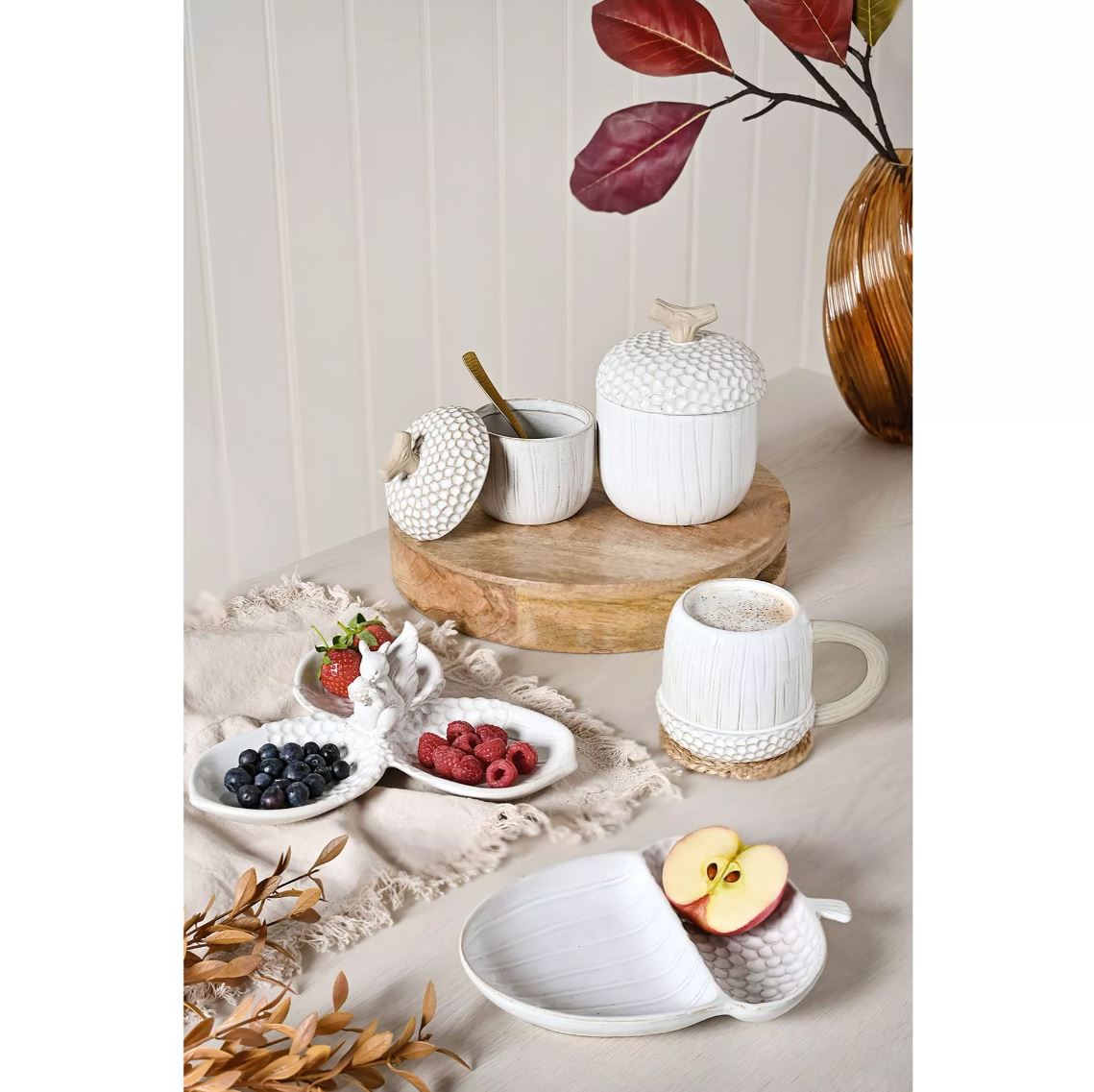 A styled tabletop arrangement of white ceramic tableware in a warm neutral setting: two acorn-shaped jars with lids, a small cup with lid, a mug with handle, a divided plate, and fruit slices on a beige cloth with dried leaves and a vase in the background.