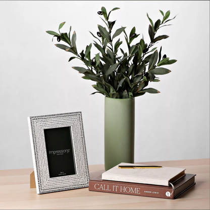 Decorative setup with a photo frame, vase with greenery, and book on a wooden surface.