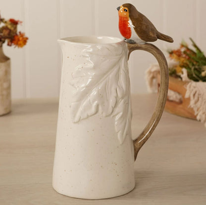 A speckled cream-colored ceramic pitcher with a raised leaf design on the front and a brown handle, featuring a small robin bird figurine perched on the rim, set on a light wooden tabletop.