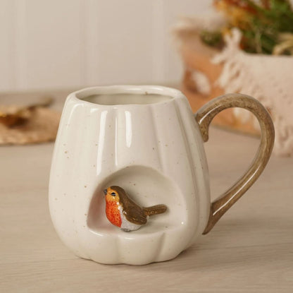 A cream-colored speckled ceramic mug with a handle, featuring a recessed niche on the front holding a small painted robin bird, set on a light wooden surface.