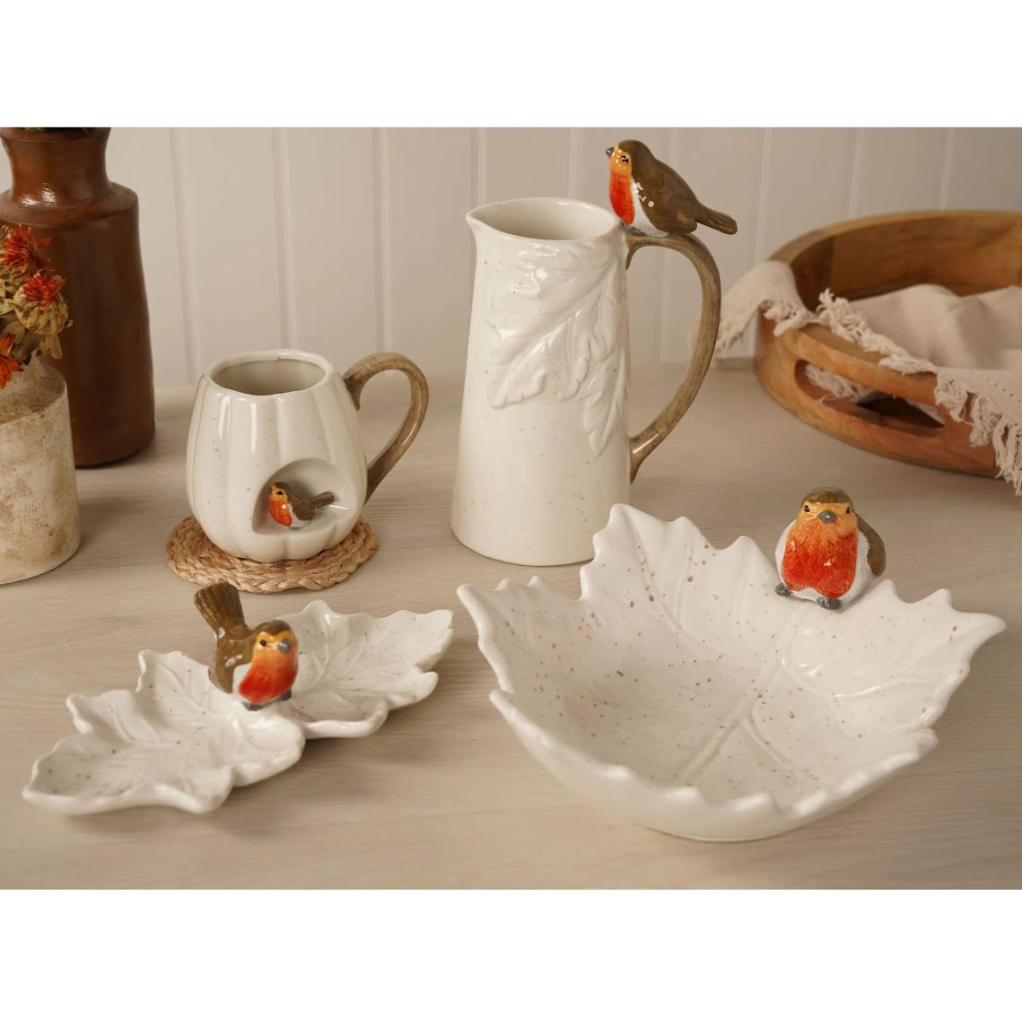 A ceramic table setting featuring a large leaf-shaped bowl, a matching leaf-shaped dish, a leaf-embossed pitcher, a small leaf-shaped cup, and several robin figurines, arranged on a light wood surface with neutral home decor props in the background.