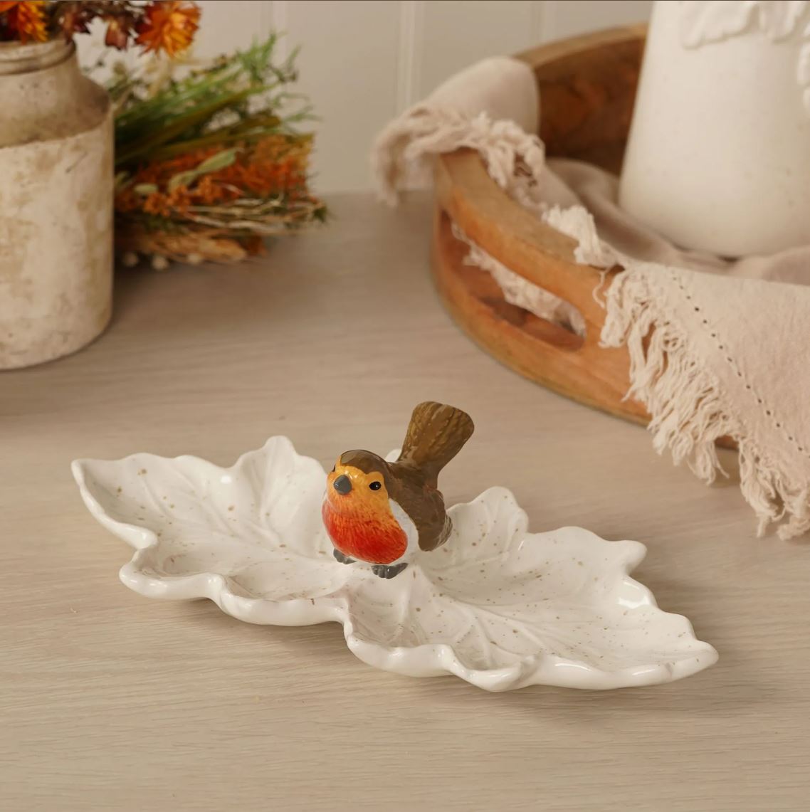A white ceramic leaf-shaped dish with a small robin bird figurine on top, placed on a light wood tabletop with softly blurred home decor in the background.