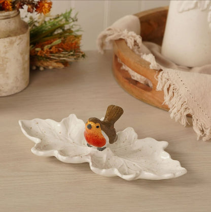 A white ceramic leaf-shaped dish with a small robin bird figurine on top, placed on a light wood tabletop with softly blurred home decor in the background.