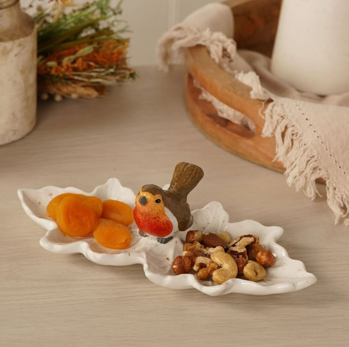 A white ceramic robin-in-a-leaf dish holding dried apricots and mixed nuts, styled on a light wood tabletop with rustic decorative props in the background.