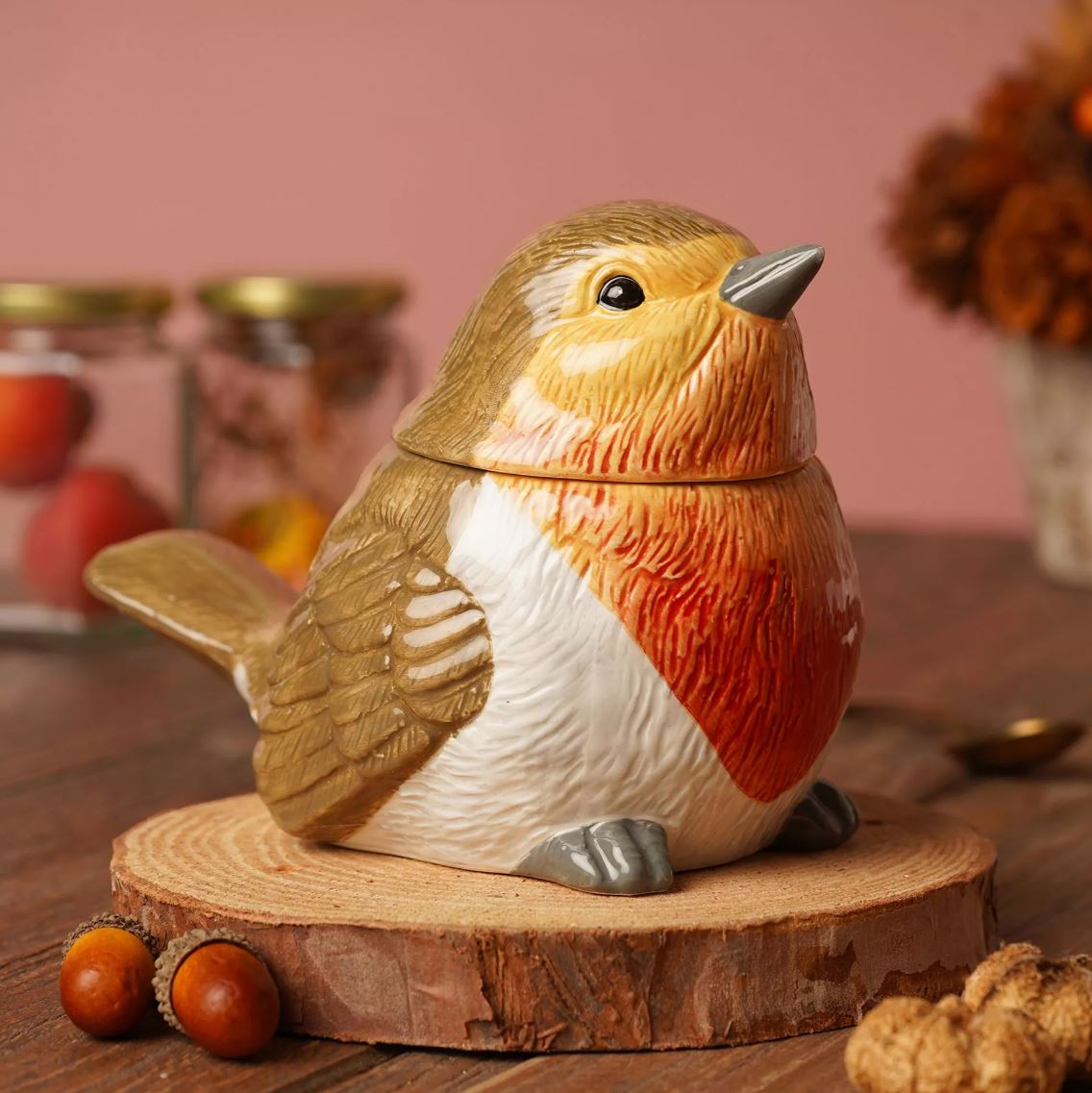 A ceramic robin bird-shaped container with a lid, displayed on a rustic wood slice coaster. The bird features a golden-brown back, a white belly, and a warm orange-red chest.