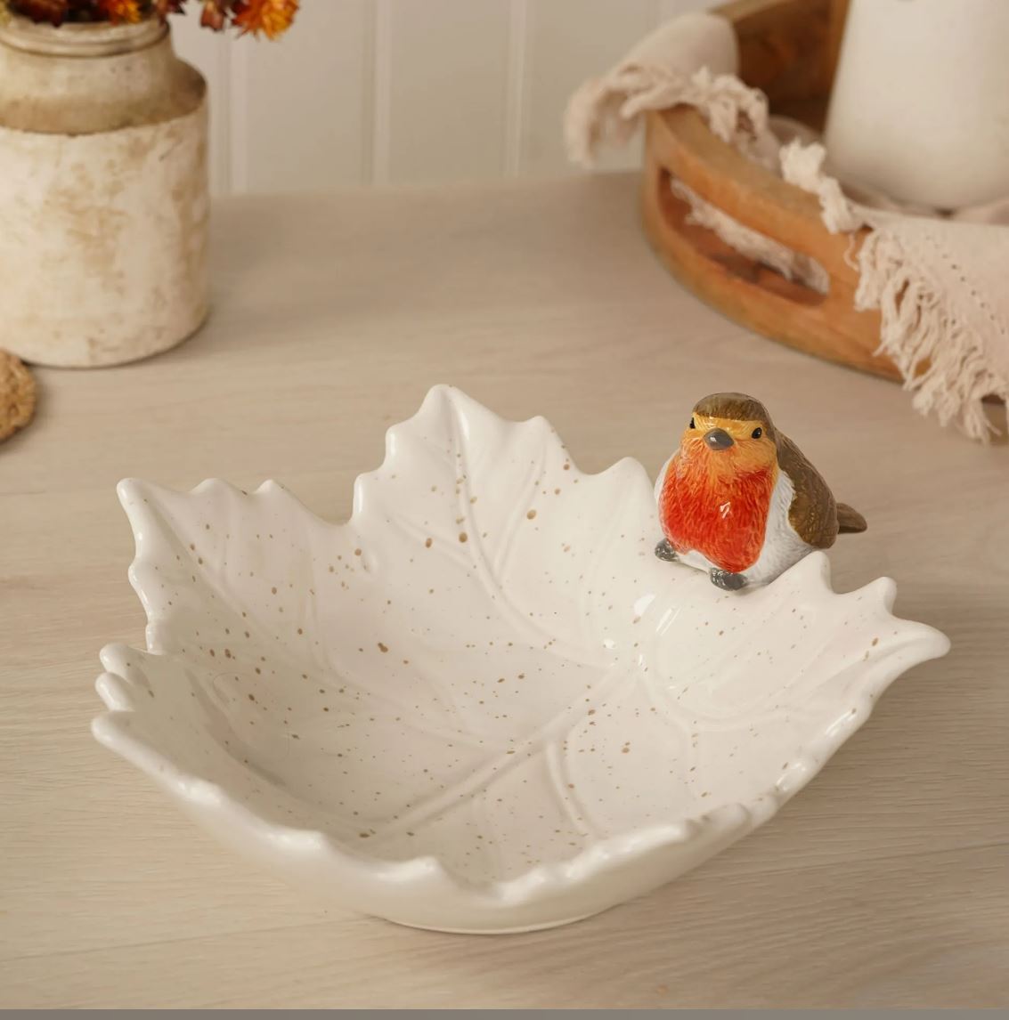 A white ceramic leaf-shaped bowl with subtle speckles, styled on a light wood tabletop with a small robin bird figurine perched on the rim.