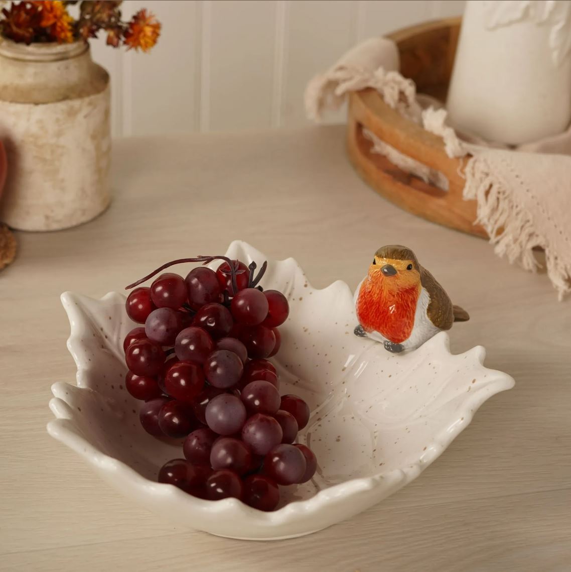 A white ceramic leaf-shaped bowl with speckled detailing, filled with dark red grapes and decorated with a small robin figurine, set on a light wood tabletop with rustic home decor in the background.