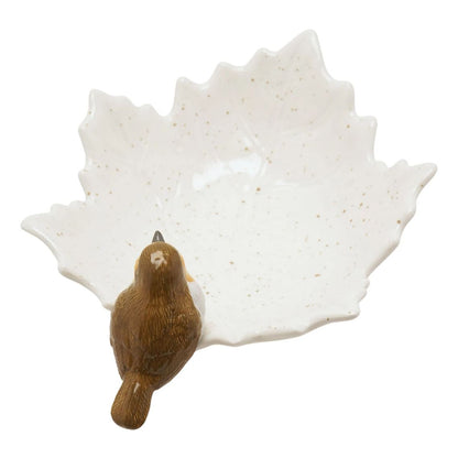 A ceramic maple leaf-shaped dish in off-white with subtle speckles, featuring a small brown bird figurine perched on the lower left edge, shown against a plain white background.