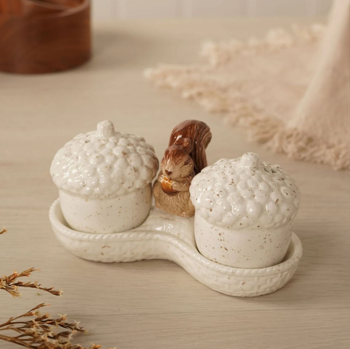 A ceramic acorn-shaped salt and pepper shaker set on a matching tray, featuring two speckled off-white acorns and a small brown squirrel figurine in the center, styled on a light wood surface.