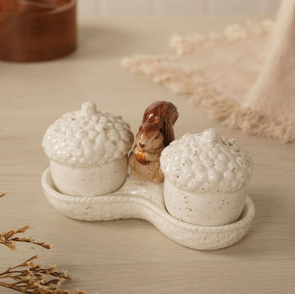A ceramic acorn-shaped salt and pepper shaker set on a matching tray, featuring two speckled off-white acorns and a small brown squirrel figurine in the center, styled on a light wood surface.
