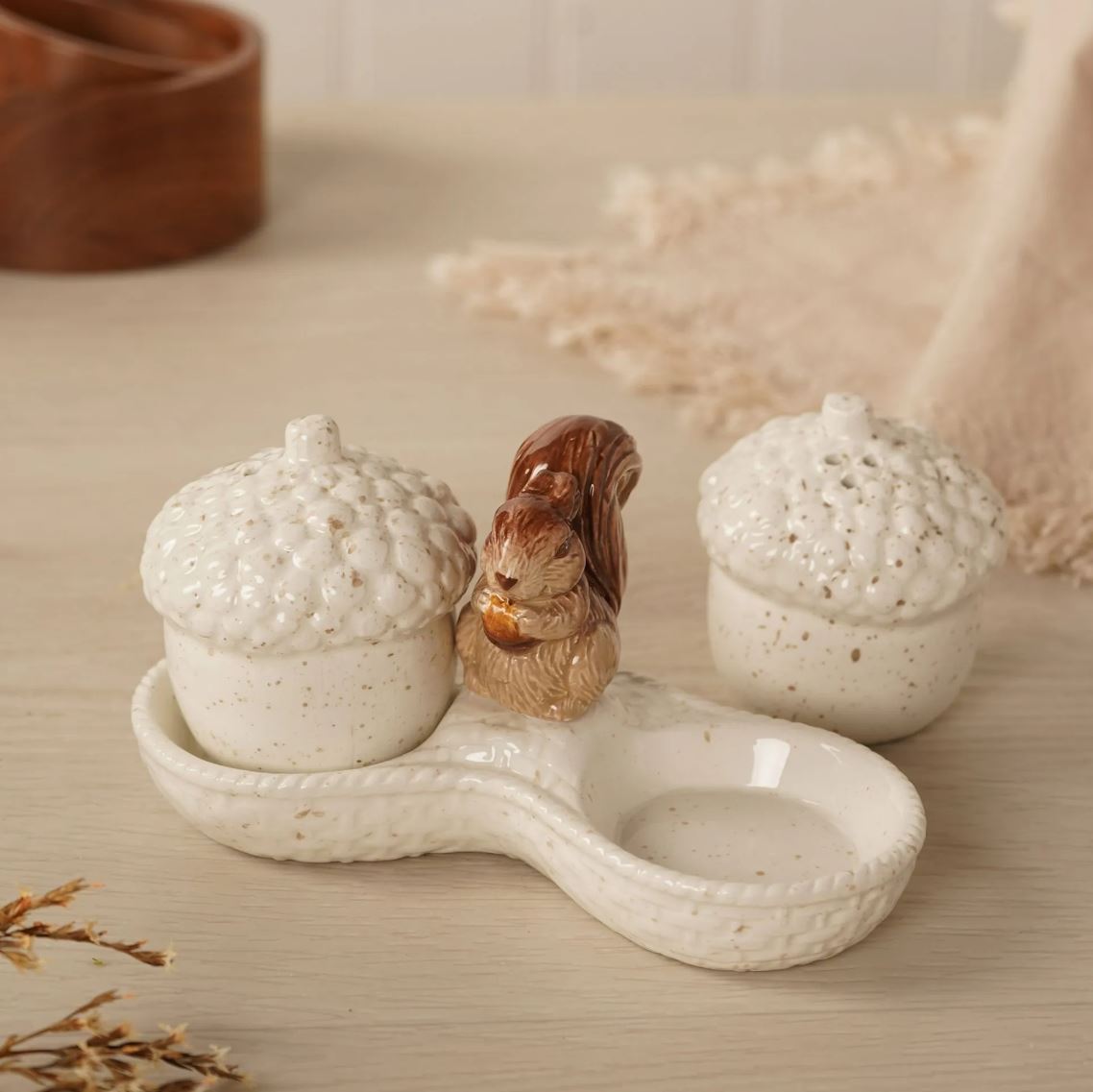 A ceramic acorn-shaped dish with two speckled cream-colored acorn lids and a small brown squirrel figurine standing in the center, set on a light wood surface with a softly blurred background.