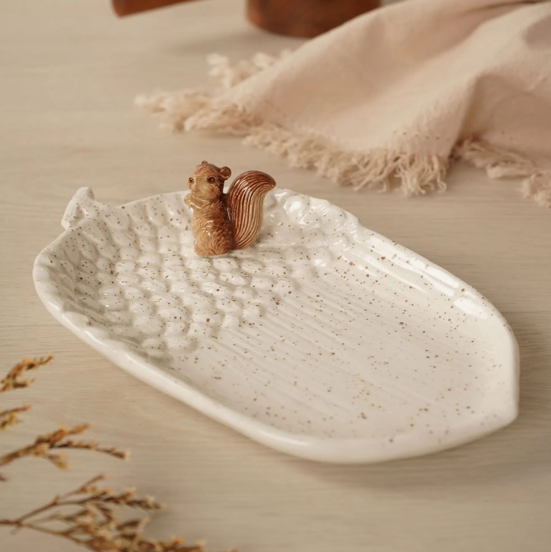 A white ceramic leaf-shaped dish with brown speckles and a small wooden squirrel figurine on top, displayed on a light wood surface with a beige fringed cloth in the background.