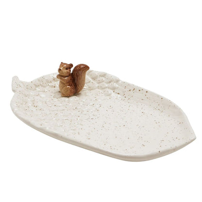 A leaf-shaped ceramic dish in an off-white glaze with brown speckles, featuring a small brown squirrel figurine on top, set against a plain white background.
