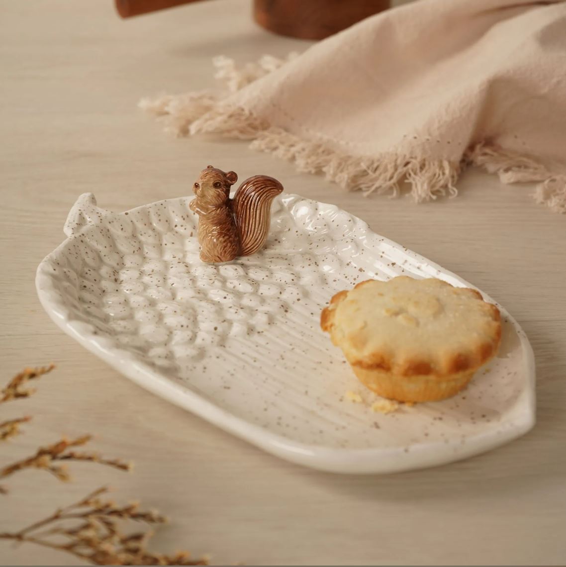A white speckled ceramic leaf-shaped serving plate with a small brown squirrel figurine and a mini tart, styled on a light wood surface with a beige fringed cloth in the background.