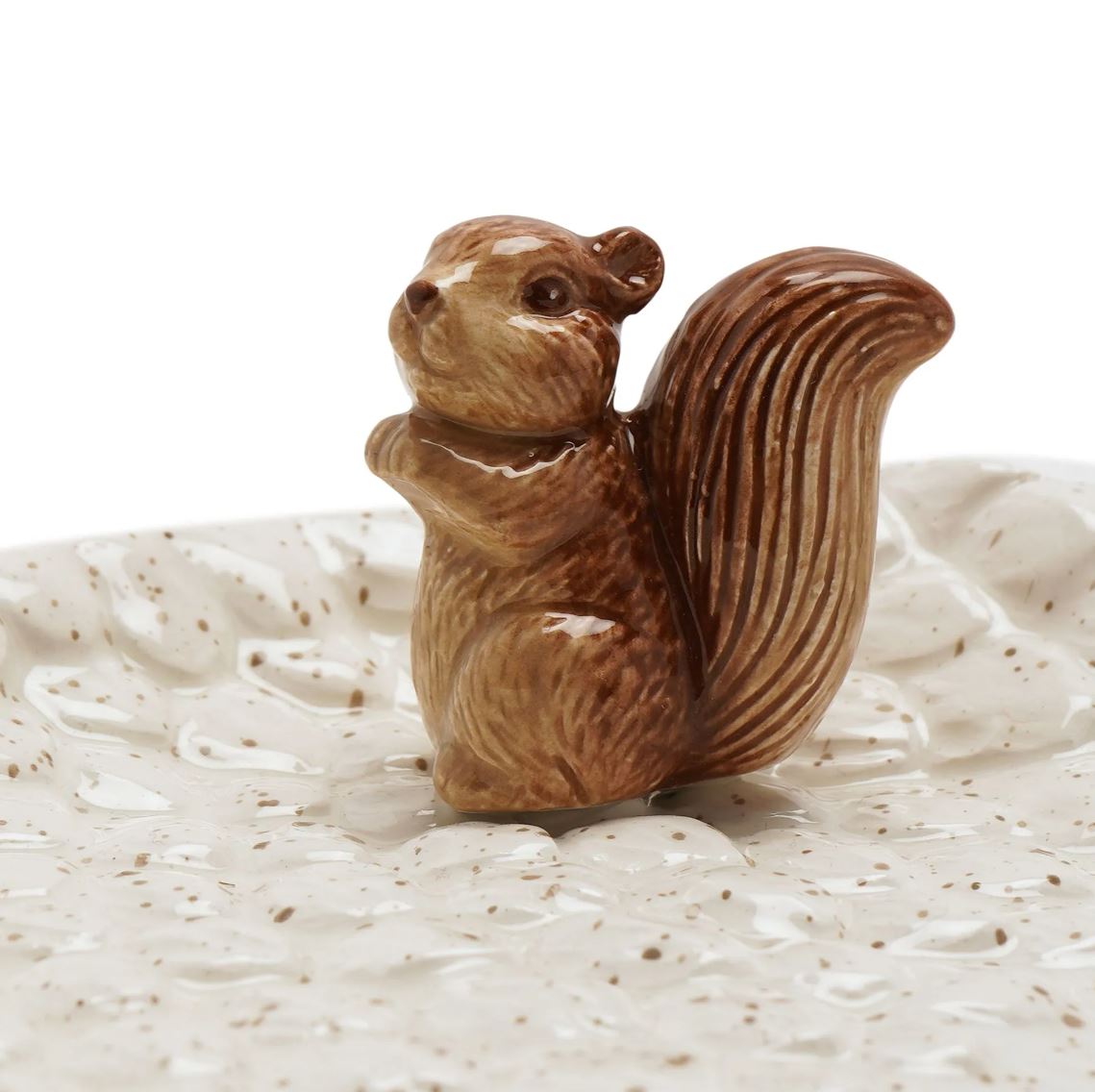 A small glazed ceramic squirrel figurine in warm brown tones, standing on a textured speckled cream dish against a white background.