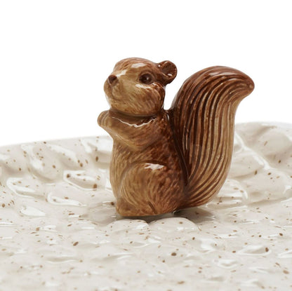 A small glazed ceramic squirrel figurine in warm brown tones, standing on a textured speckled cream dish against a white background.