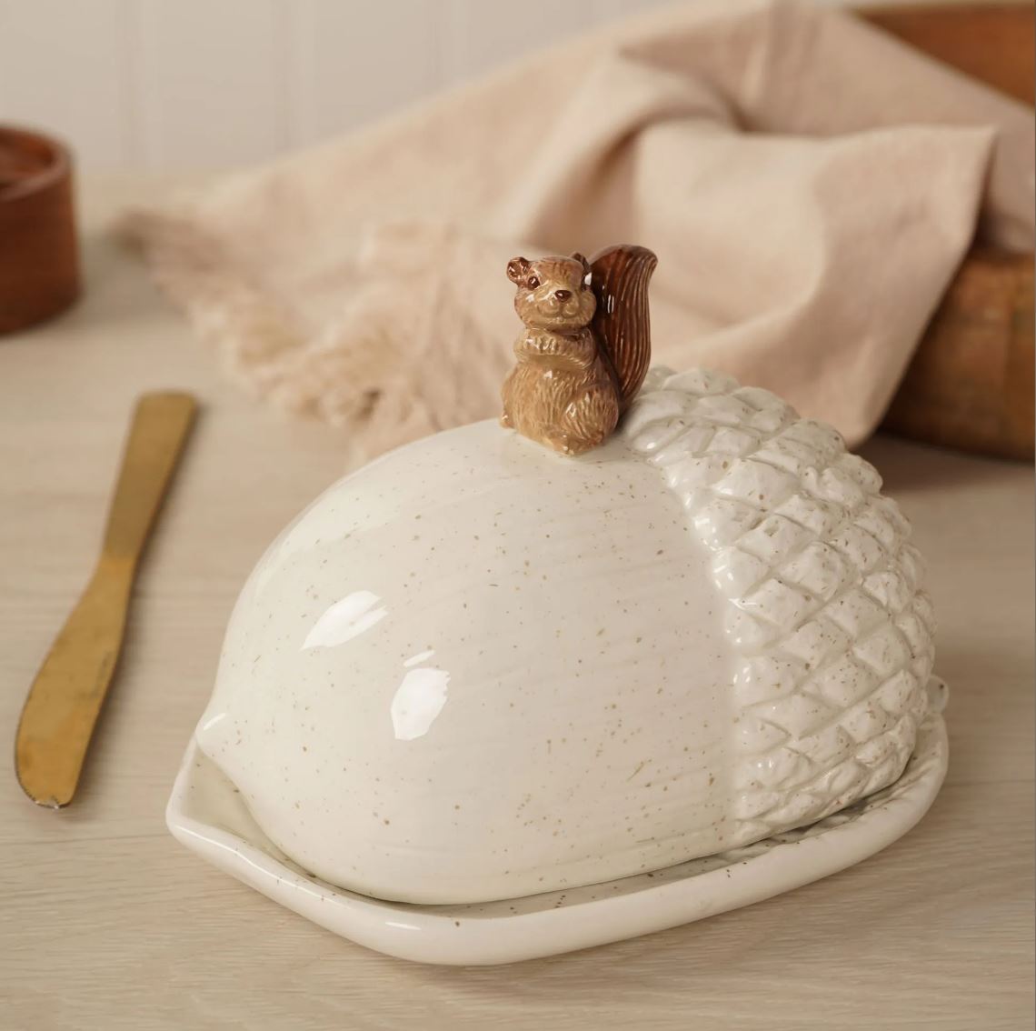 A ceramic butter dish shaped like an acorn with a small carved squirrel figurine on top, shown on a tabletop with a napkin and wooden spoon in a softly styled setting.