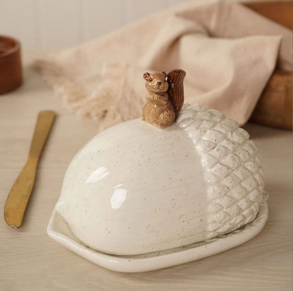 A ceramic butter dish shaped like an acorn with a small carved squirrel figurine on top, shown on a tabletop with a napkin and wooden spoon in a softly styled setting.