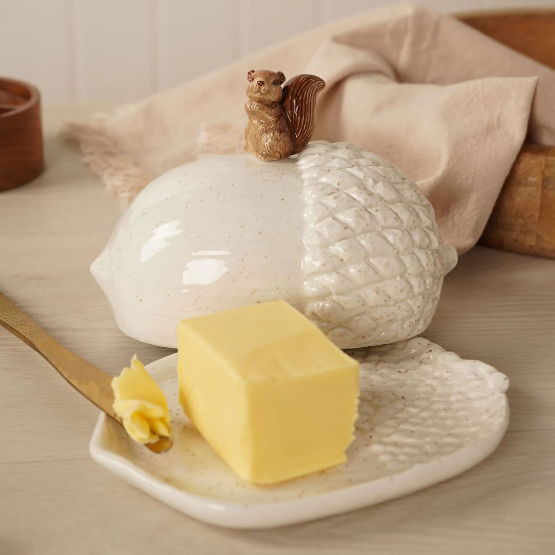 A ceramic acorn-shaped butter dish with a small squirrel figurine on top, shown with a stick of butter on a matching ceramic plate in a warm, styled kitchen setting.