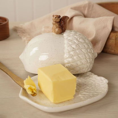 A ceramic acorn-shaped butter dish with a small squirrel figurine on top, shown with a stick of butter on a matching ceramic plate in a warm, styled kitchen setting.