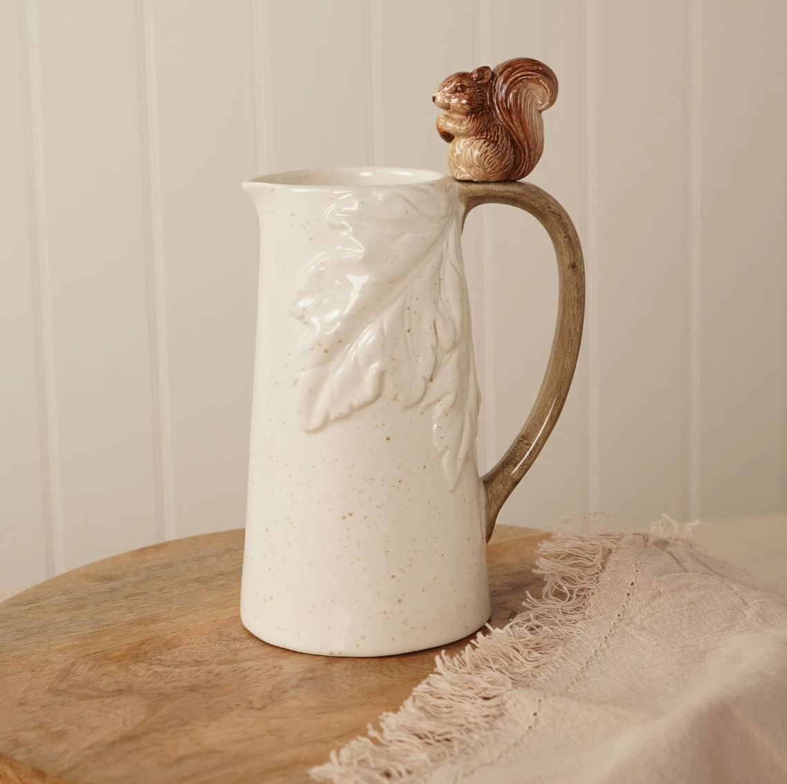A speckled cream ceramic pitcher with a raised leaf design on the side, a brown handle, and a small carved squirrel figure on top, set on a wooden surface with a fringed textile nearby.