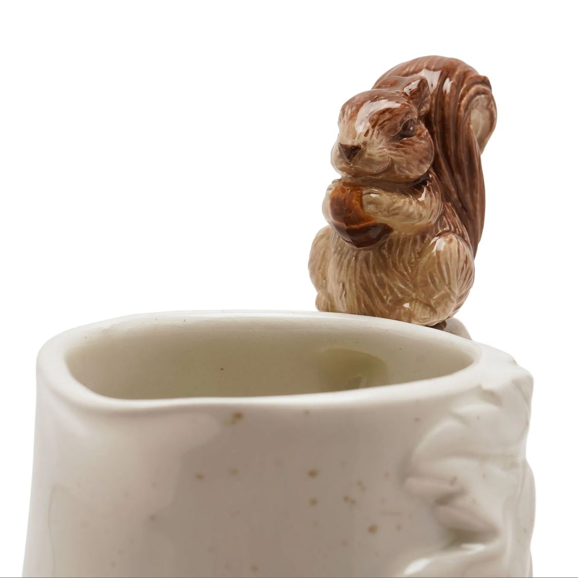 A ceramic mug with a brown glazed squirrel figurine holding an acorn perched on the rim, shown against a plain white background.