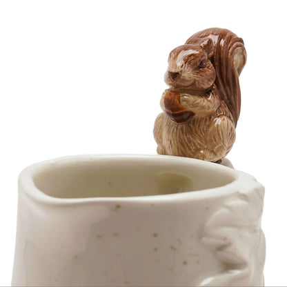 A ceramic mug with a brown glazed squirrel figurine holding an acorn perched on the rim, shown against a plain white background.
