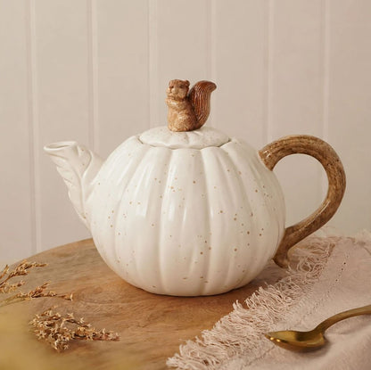 A white ceramic pumpkin-shaped teapot with subtle speckles, featuring a small brown squirrel figurine on top of the lid and a rustic brown handle, displayed on a wooden surface with a gold spoon and dried flowers.