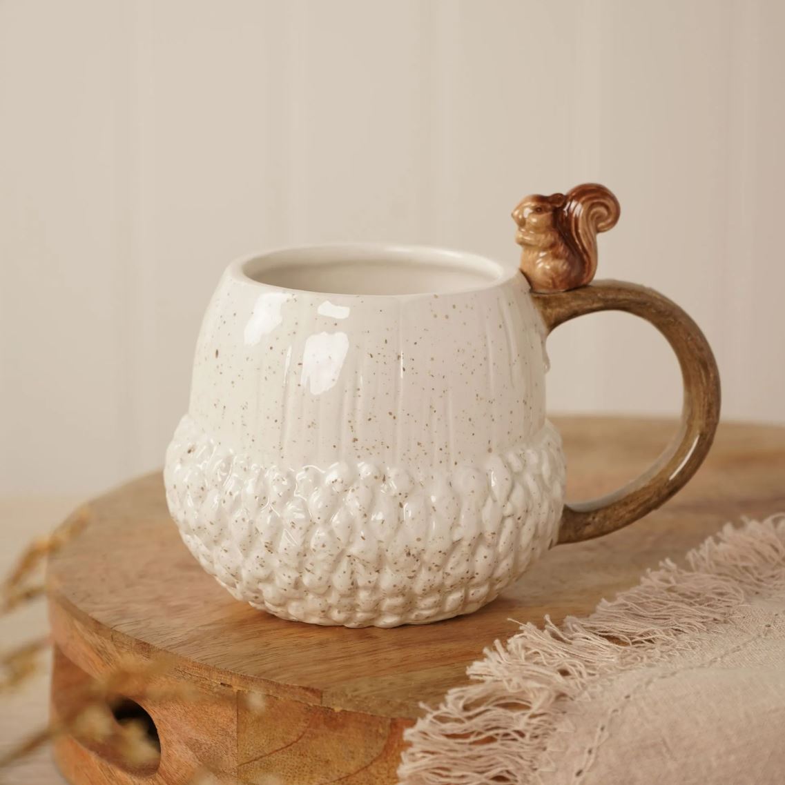 A ceramic mug designed to look like an acorn, featuring a textured cream-colored body with brown speckles, a brown handle, and a small 3D squirrel ornament on top, displayed on a wooden board.