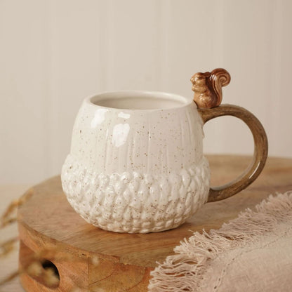 A ceramic mug designed to look like an acorn, featuring a textured cream-colored body with brown speckles, a brown handle, and a small 3D squirrel ornament on top, displayed on a wooden board.