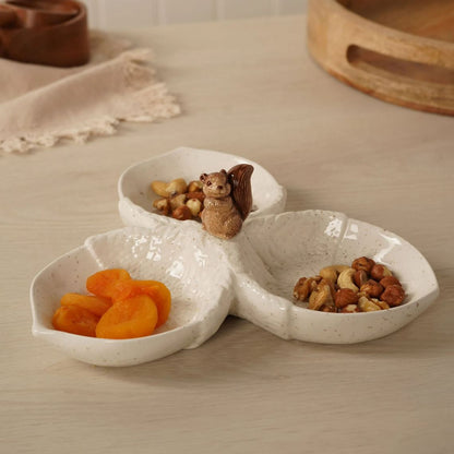 A three-compartment ceramic snack plate with a textured, speckled off-white finish, styled on a light wood table. The compartments are filled with dried apricots, nuts, and a small decorative squirrel figurine sits on the central junction.