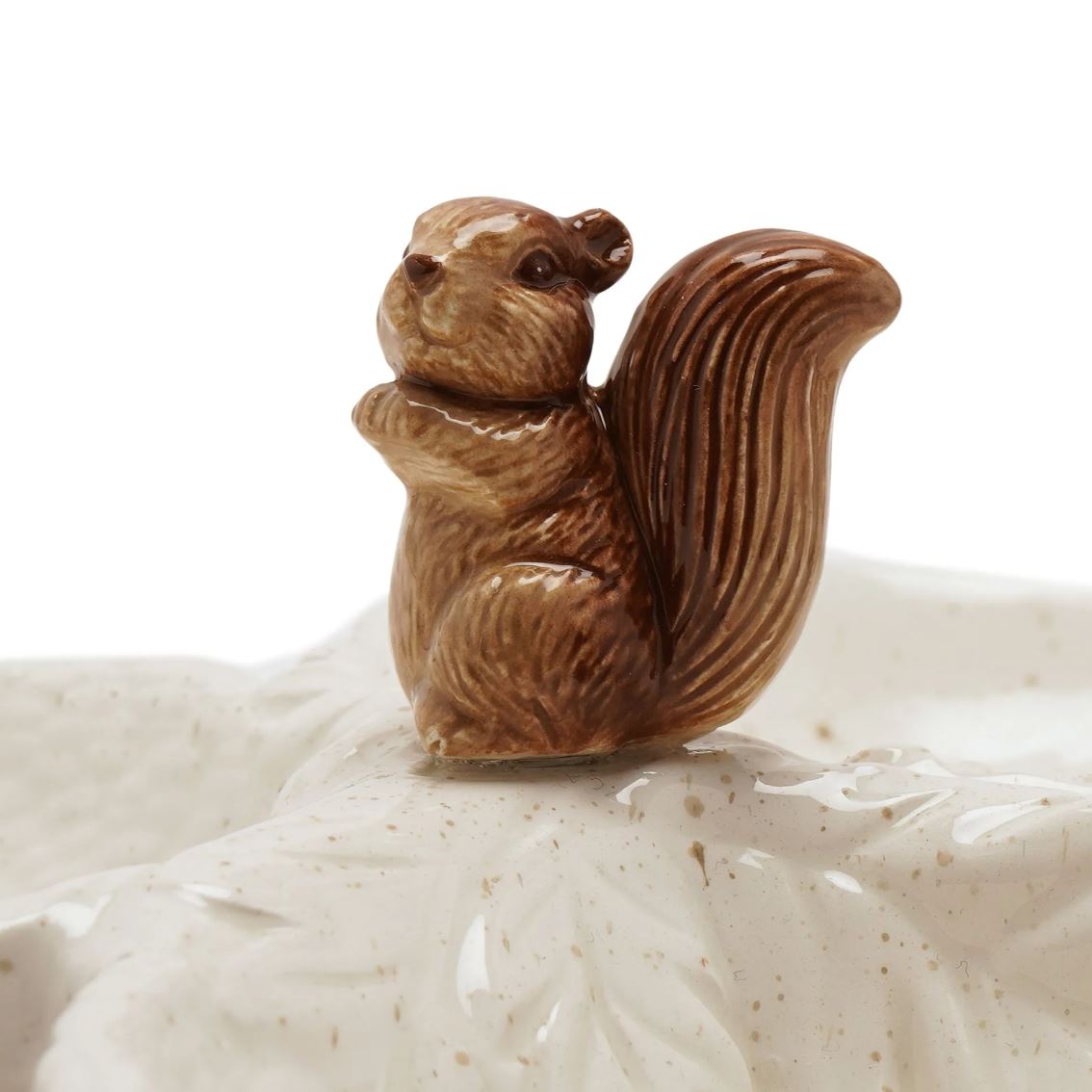 A small glazed ceramic squirrel figurine in warm brown tones, standing on a white speckled ceramic surface against a plain white background.