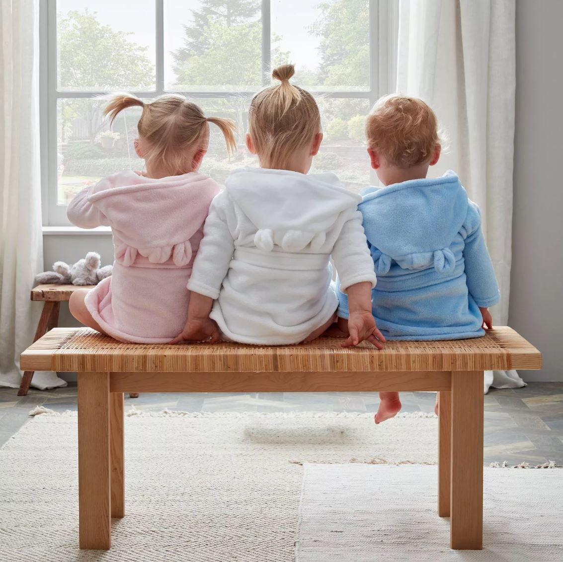 Three toddlers wearing pink, white, and blue hooded robes sit on a wooden bench, facing a large window in a bright room.