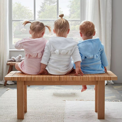 Three toddlers wearing pink, white, and blue hooded robes sit on a wooden bench, facing a large window in a bright room.