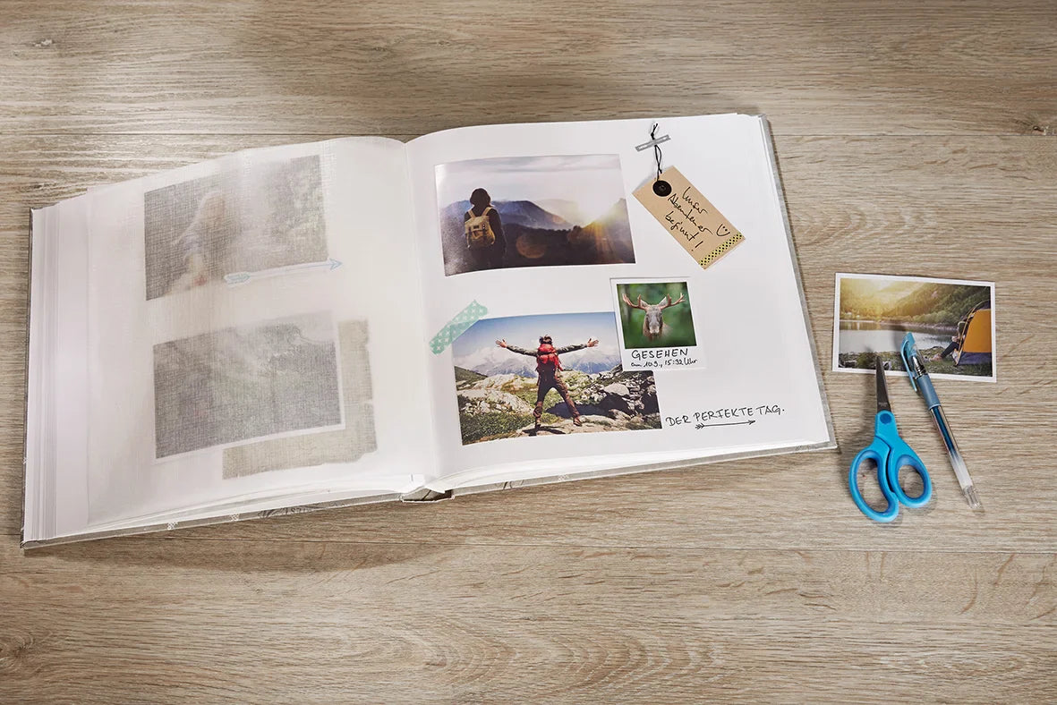 Open photo album with scenic photos on a wooden surface, accompanied by scissors and a pen.
