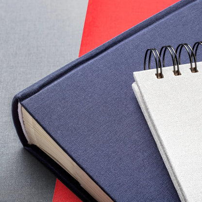 A close-up of a navy blue photo album with a white page and black spiral binding, placed on top of another red book against a grey textured background.