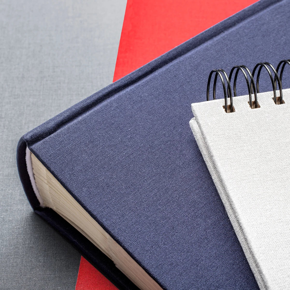 A close-up of a navy blue photo album with a white page and black spiral binding, placed on top of another red book against a grey textured background.