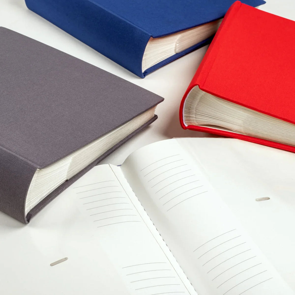 Three hardcover albums with blue, red, and grey covers arranged on a white surface, with an open white album in the foreground.
