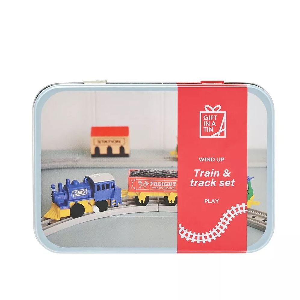 A toy train and track set inside a clear rectangular gift tin with a red front label that reads 'Gift in a Tin' and 'Play'. The set includes a blue and yellow locomotive, a red and yellow freight car, and a small station figure on a gray track base.